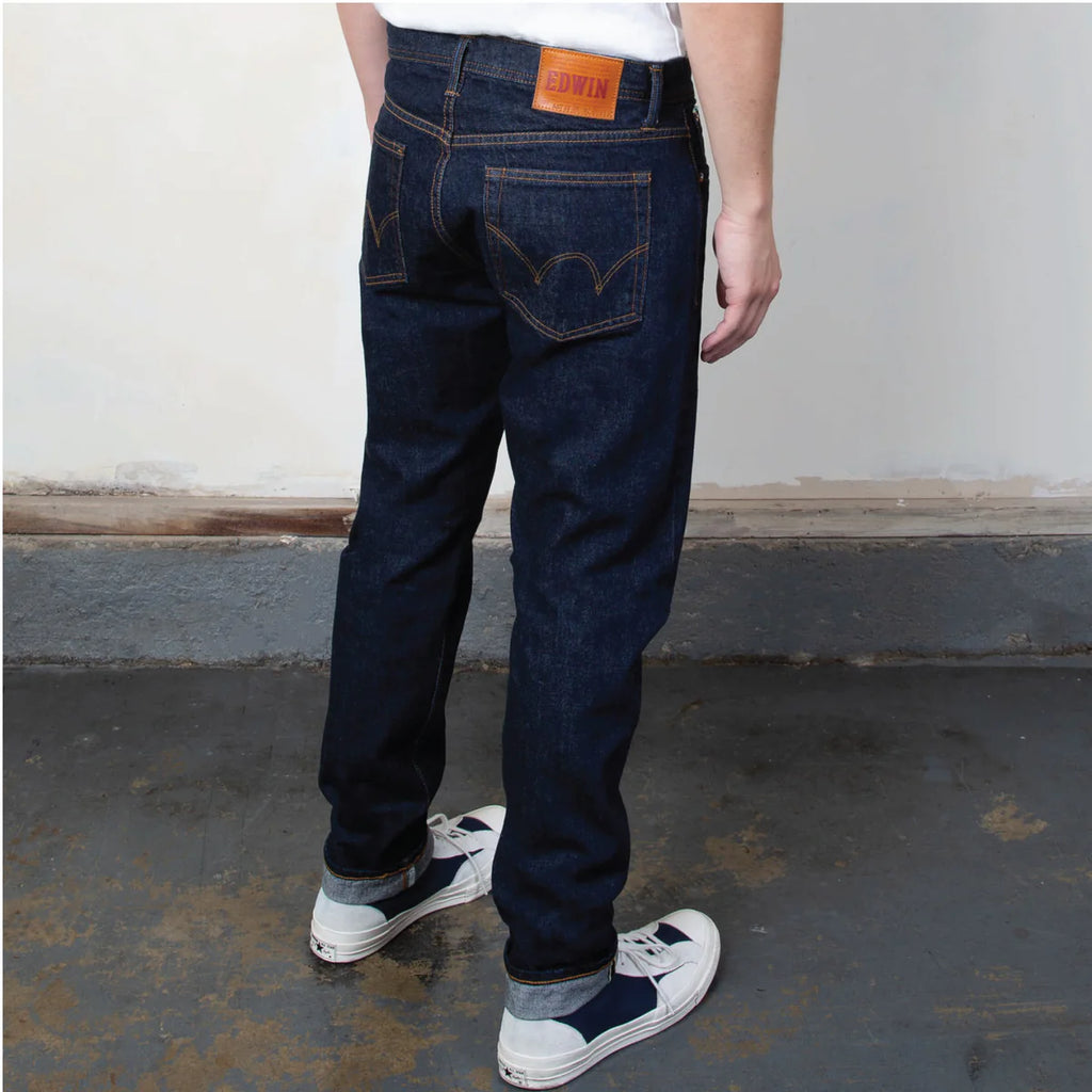 Person wearing dark blue jeans with an orange brand label on a concrete floor.