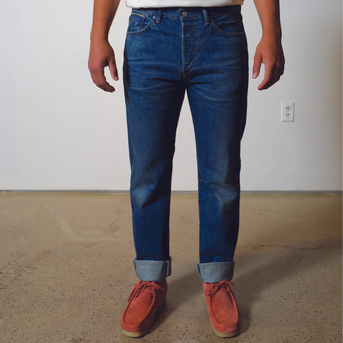 Person wearing blue jeans and red shoes standing on a concrete floor with a white wall background.
