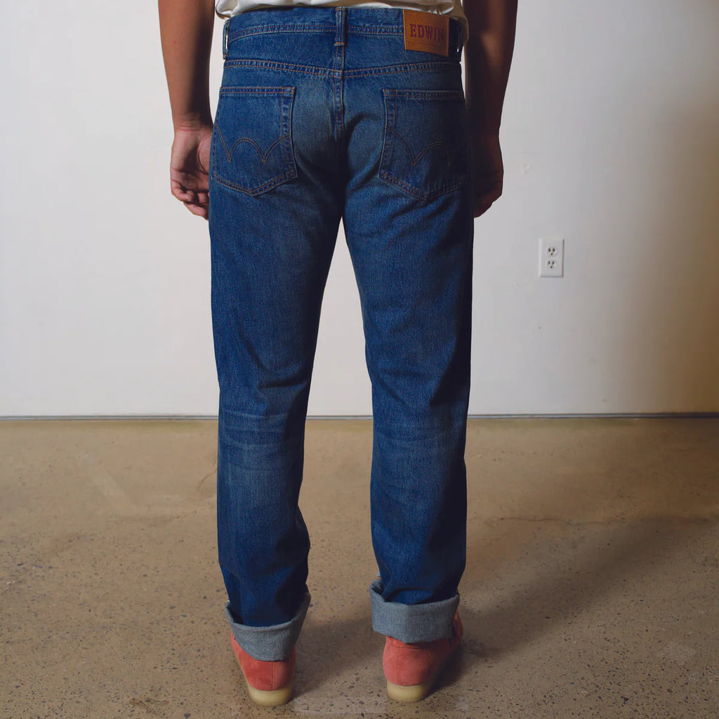 Person wearing blue jeans standing in a room with a plain wall and carpeted floor.